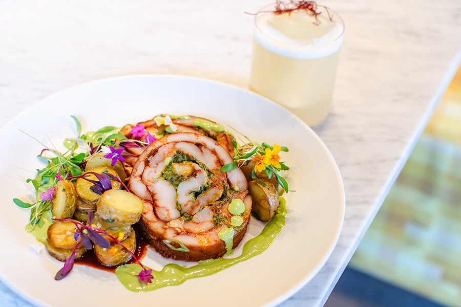 Rolled and sliced chicken roulade dish garnished with green sauce, microgreens, edible flowers, and roasted baby potatoes, served on a white plate. A light-colored cocktail with foam and a garnish sits in the background on a marble surface.