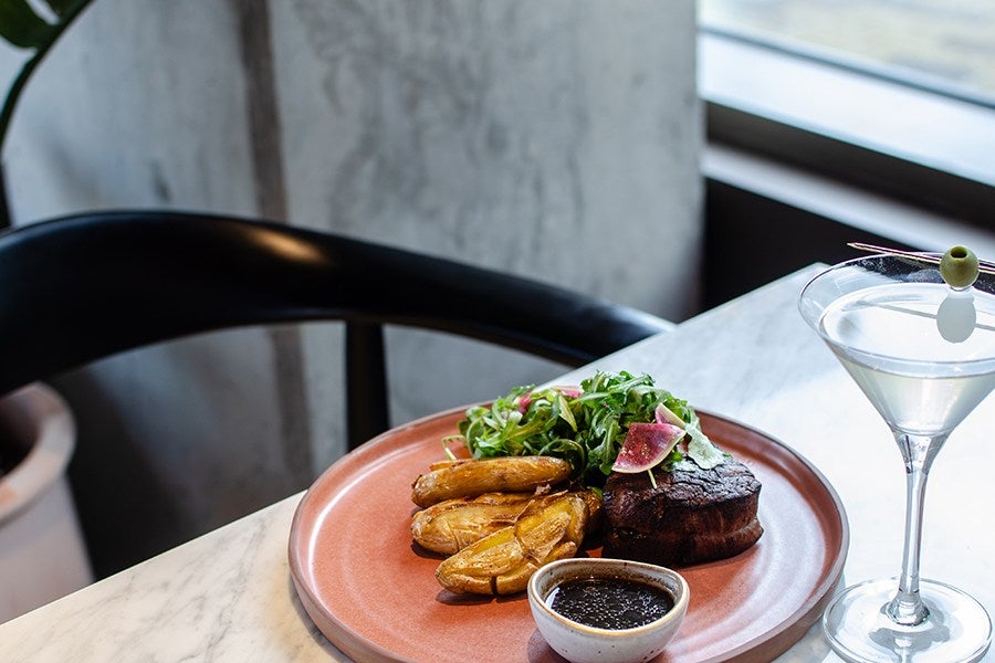 Grilled steak served with roasted fingerling potatoes and a fresh green salad with radish slices on a terracotta plate, accompanied by a small bowl of dark sauce. A martini glass with a clear cocktail garnished with a green olive on a toothpick is placed next to the plate on a marble table.