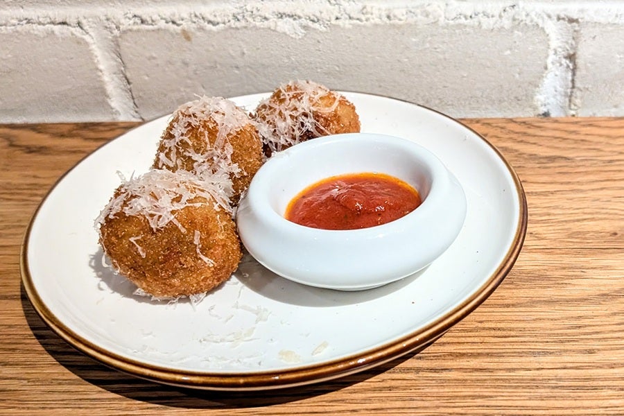 Three breaded and fried arancini balls topped with grated cheese, served on a white plate with a small white bowl of marinara sauce, placed on a wooden surface with a white brick wall background.