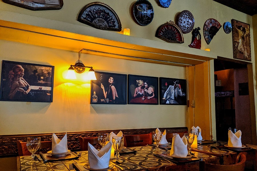A dining table set for six with folded white napkins, wine glasses, and plates on a patterned tablecloth. The wall behind the table features four framed paintings of people, a wall-mounted light fixture with two lamps, and decorative fans and plates arranged above. The room has warm yellow lighting and wooden chairs around the table.
