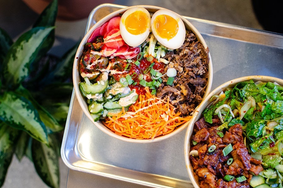 Two bowls of food on a metal tray, one bowl contains soft-boiled eggs, shredded carrots, sliced cucumbers, pickled vegetables, cooked beef, and green onions with sauce drizzled on top; the other bowl contains cooked meat, leafy greens, and sliced cucumbers with green onions sprinkled over.