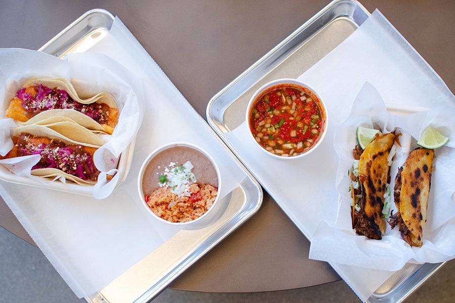 Two trays each with two tacos wrapped in paper. The left tray has tacos with fried fish and purple cabbage, accompanied by a cup of refried beans and Mexican rice. The right tray has tacos with grilled meat and onions, served with a cup of salsa and lime wedges.