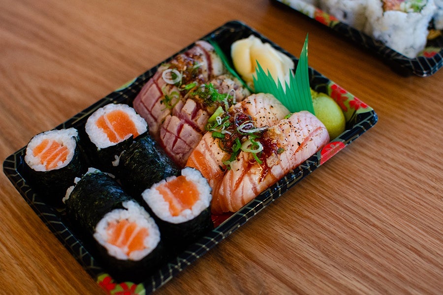 A black plastic tray containing an assortment of sushi, including salmon maki rolls wrapped in seaweed, and nigiri topped with seared salmon and garnished with green onions and sauce. The tray also includes pickled ginger and a small portion of wasabi, with a decorative green plastic leaf divider. The tray is placed on a wooden surface.
