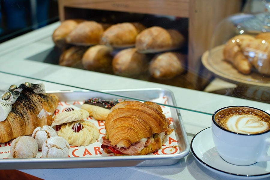 A metal tray holds a variety of pastries including a croissant sandwich with ham and cheese, a croissant topped with toasted marshmallows, several small powdered sugar cookies, and a cookie with chocolate sprinkles. Next to the tray is a white cup of cappuccino with latte art on top, placed on a matching saucer. In the background, there is a display case with more croissants and bread.