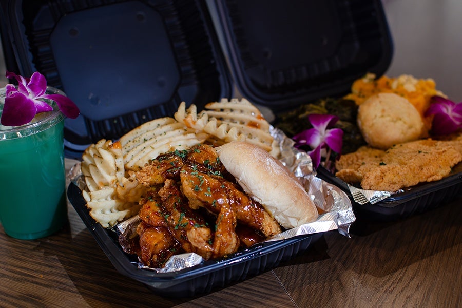 Two black takeout containers with food are shown. The front container holds waffle fries, several pieces of sauced fried chicken, and a sandwich bun. The back container contains a breaded fried fish fillet, a biscuit, cooked greens, mashed sweet potatoes, and is garnished with purple orchid flowers. A blue-green drink with a purple orchid flower on top is placed to the left of the containers.