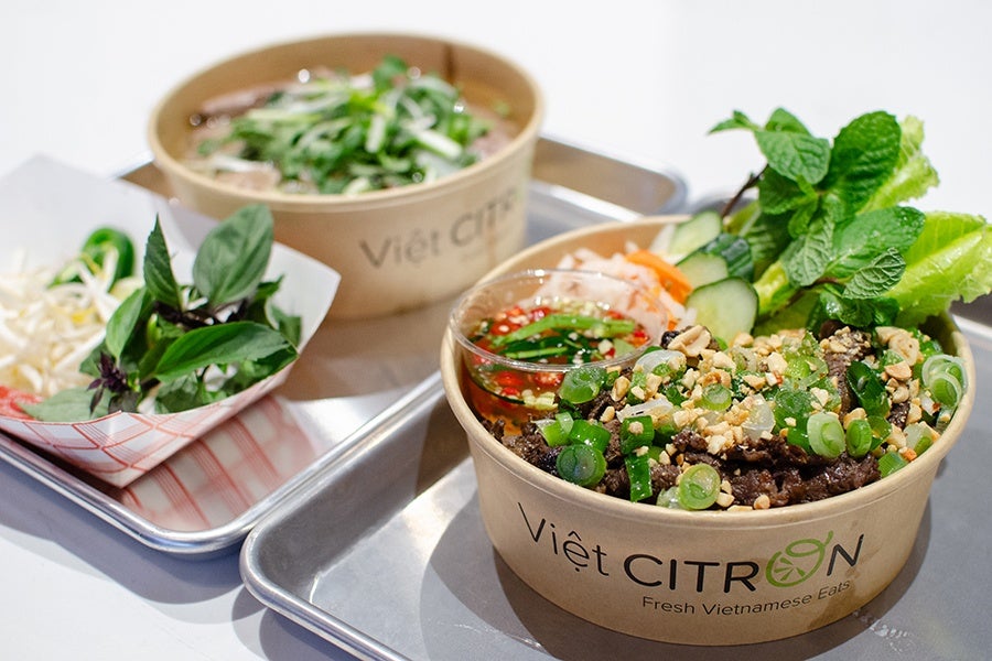 A bowl of Vietnamese food topped with chopped green onions, crushed peanuts, and fresh herbs, accompanied by a small dish of dipping sauce, sliced cucumbers, and lettuce. In the background, there is another bowl of Vietnamese soup garnished with herbs, and a paper tray containing bean sprouts and fresh basil leaves. Both bowls are placed on metal trays, and the front bowl has the text "Việt CITRON Fresh Vietnamese Eats" printed on it.