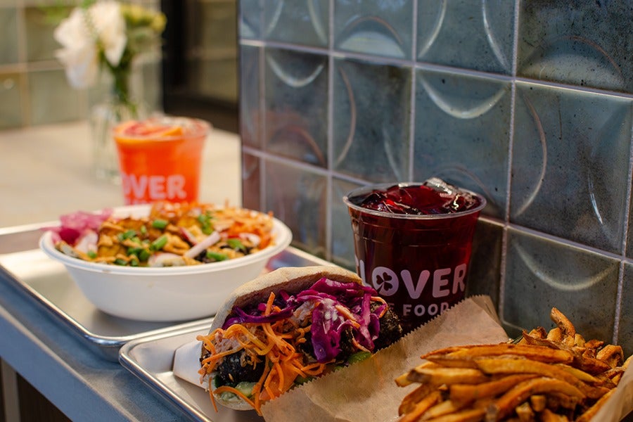 A tray with a pita sandwich filled with shredded carrots, purple cabbage, and other vegetables, a cup of dark iced beverage with ice cubes, and a serving of golden-brown fries. In the background, there is a bowl of salad with grilled chicken and a bright orange drink with a lemon slice, both slightly out of focus. The setting includes a tiled wall with circular patterns.