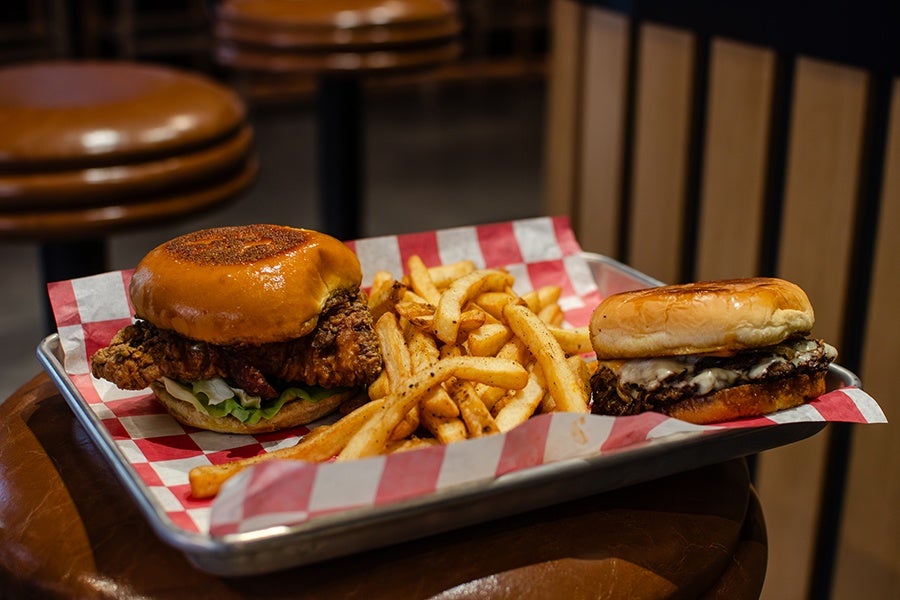A metal tray lined with red and white checkered paper holds two sandwiches and a serving of seasoned French fries. On the left is a fried chicken sandwich with a glossy toasted bun, lettuce, and crispy fried chicken. On the right is a cheeseburger with melted cheese and a toasted bun. The tray is placed on a round wooden table with brown cushioned stools in the background.
