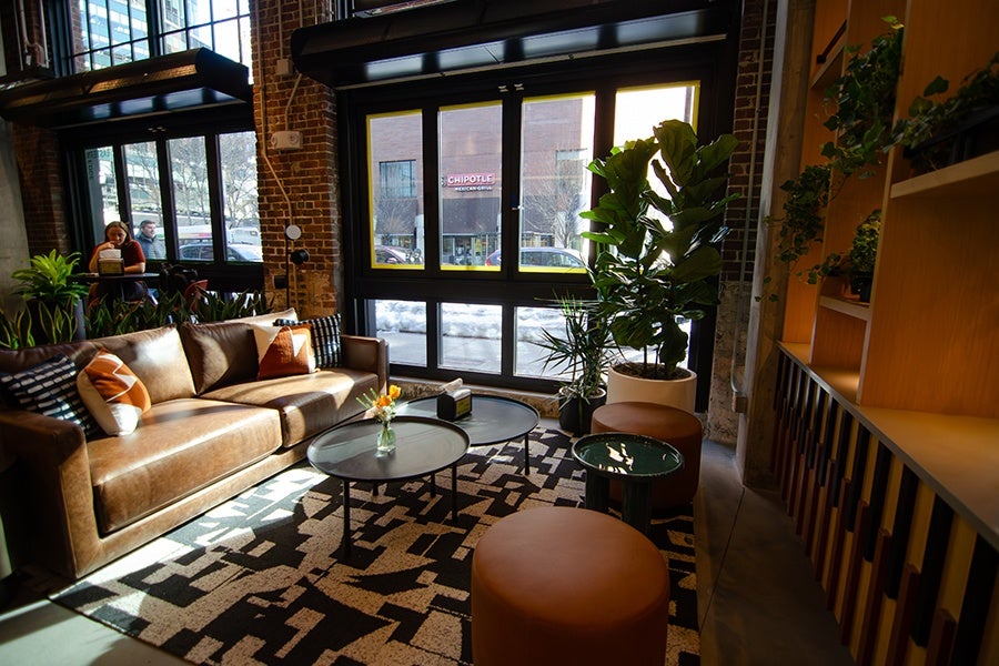 A cozy seating area with a brown leather sofa adorned with patterned cushions, two round coffee tables with a small vase of flowers, and two round brown ottomans. The space features large windows letting in natural light, a patterned rug, several green plants, and wooden shelving with additional greenery on the right side.
