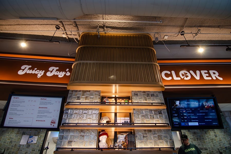 A restaurant interior featuring a large, central wooden vent hood above a tiled wall with glass block accents. Below the vent hood are shelves displaying various decorative items, including a white bust, a helmet, and small figurines. On either side of the vent hood, illuminated menu boards are visible, with the left board showing food options and the right board displaying digital content. The restaurant name "Juicy Jay's" is partially visible on the left, and "CLOVER" is prominently displayed on the right in white letters on an orange background. A person wearing a green shirt and cap stands near the right side of the image.