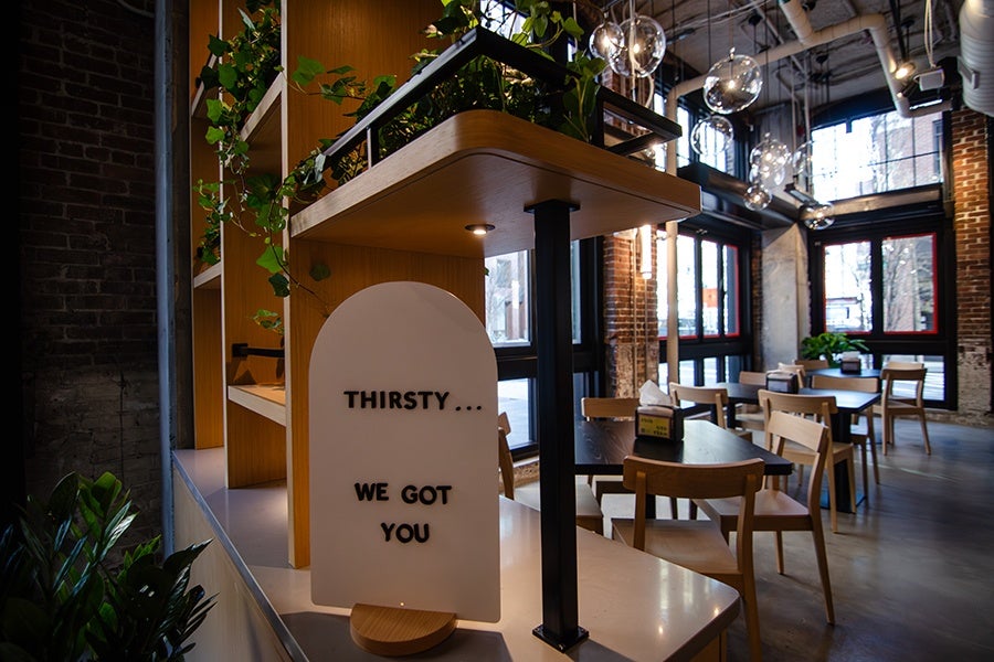 A modern café interior with wooden tables and chairs, large windows with black and red frames, and hanging globe pendant lights. In the foreground, a white sign with black text reads "THIRSTY... WE GOT YOU," placed on a counter with plants and wooden shelving. The space features exposed brick walls and an industrial ceiling with visible pipes.
