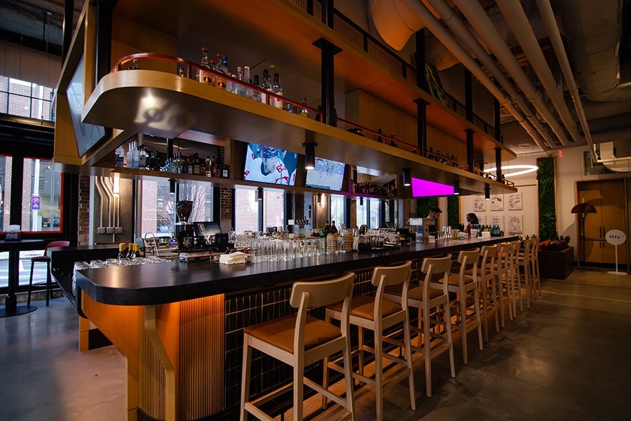 A modern bar with a long black countertop and wooden base, lined with light wooden bar stools with tan cushions. Above the bar, there is a suspended wooden shelf holding various bottles of liquor. Multiple flat-screen TVs are mounted behind the bar, displaying a hockey game. The bar area is well-lit with hanging lights, and the space features exposed pipes on the ceiling and large windows letting in natural light. The floor is polished concrete, and there is a cozy seating area with a lamp and plants in the background.