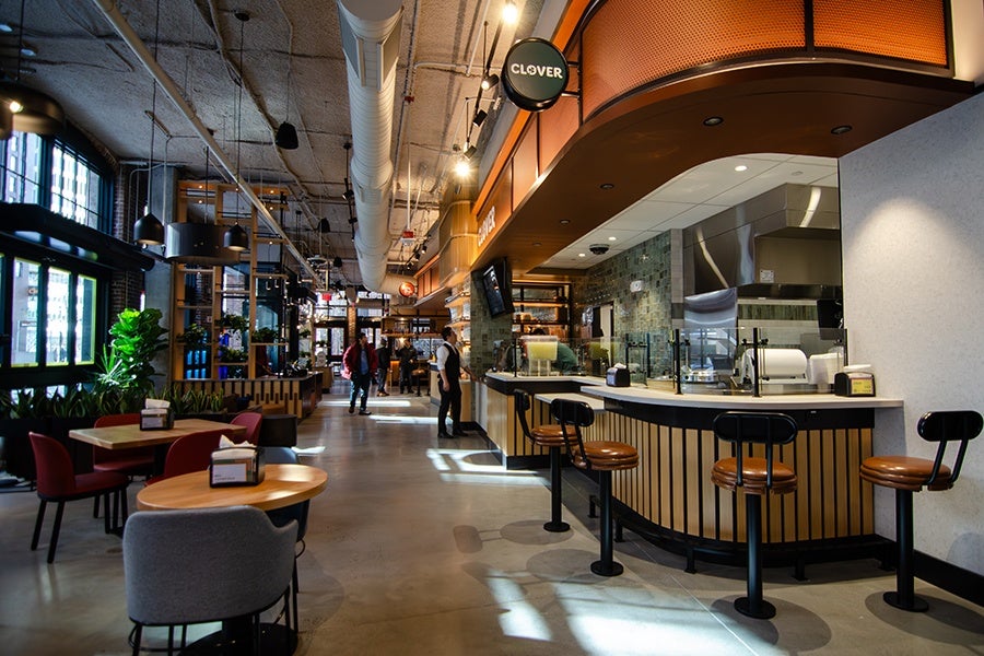 Modern cafe interior with a curved counter featuring wooden paneling and four black bar stools with brown seats. The ceiling has exposed ductwork and industrial lighting. There are tables and chairs on the left side, including red and gray upholstered chairs. Large windows on the left let in natural light, and plants add greenery near the seating area. A round sign above the counter reads "CLOVER." Several people are visible in the background near the counter and entrance.