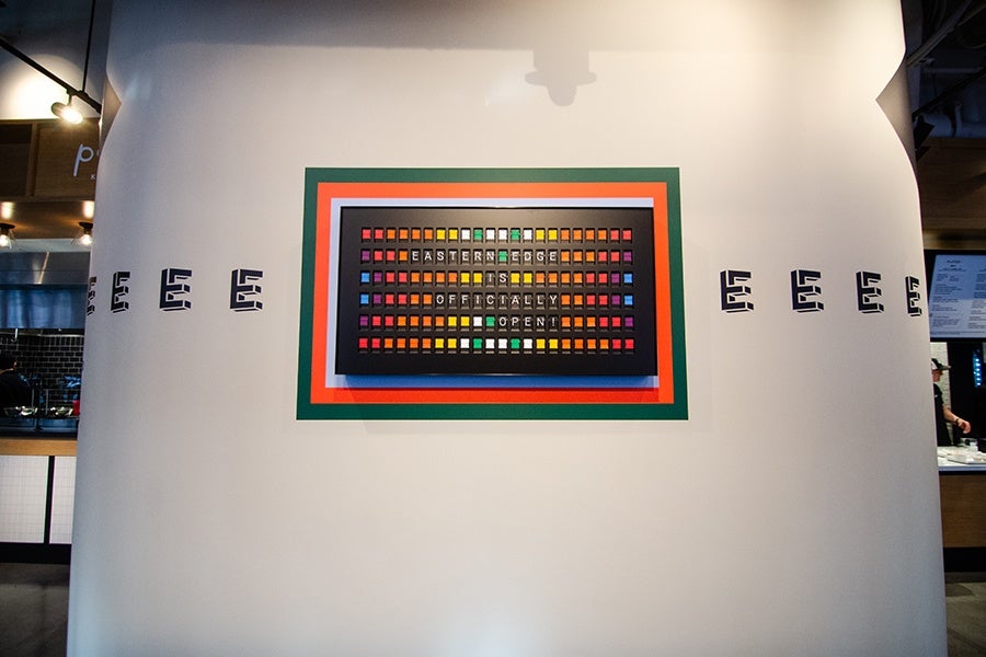 A white wall features a colorful rectangular sign with multiple small colored squares arranged in rows. The sign reads "EASTERN EDGE IS OFFICIALLY OPEN." The sign is bordered by green, orange, and white frames. On either side of the sign, the letter "E" is repeated multiple times in a stylized black font. The surrounding area appears to be an indoor space with some kitchen or restaurant elements visible on the left and right edges.
