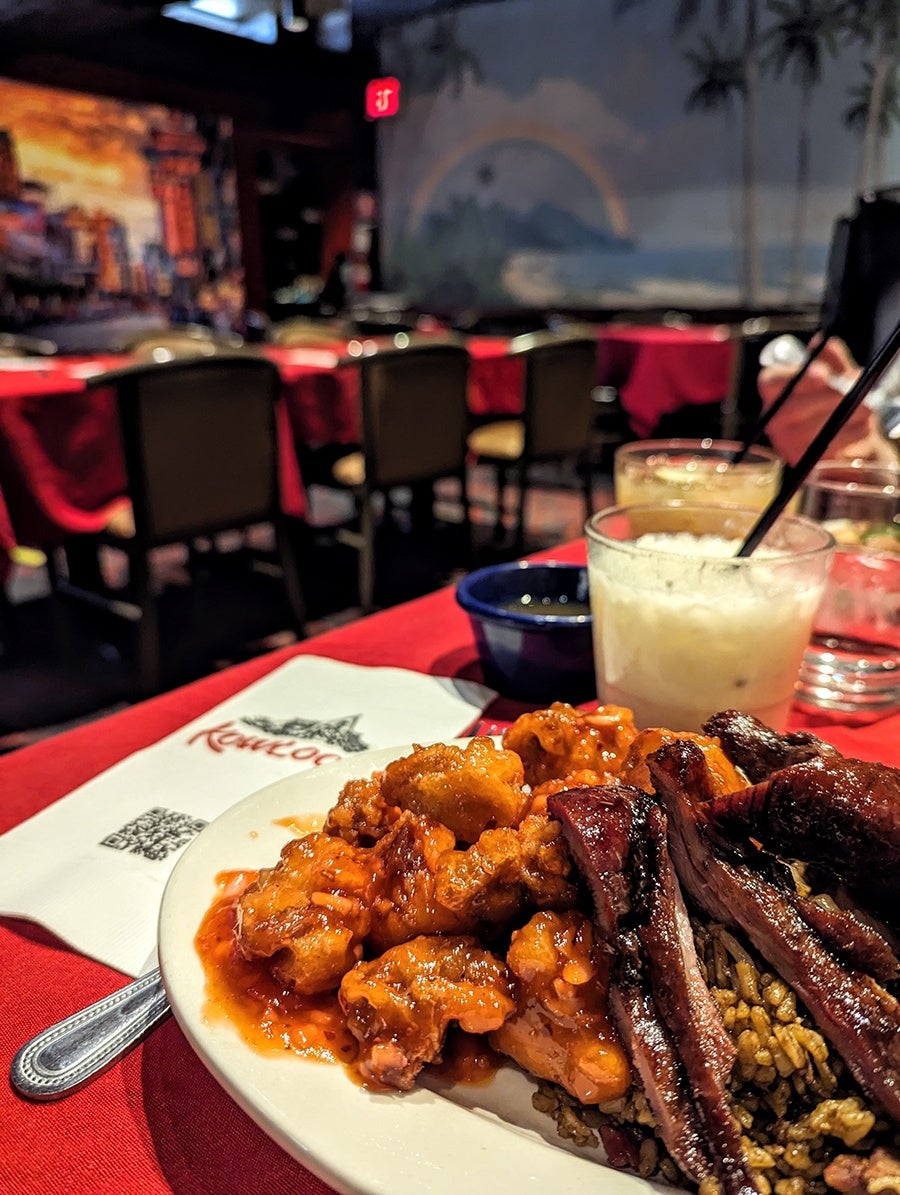 A plate of food featuring crispy fried chicken pieces coated in a reddish sauce, accompanied by slices of grilled meat on a bed of seasoned rice. The plate is on a red tablecloth with a napkin and a spoon nearby. In the background, there are two drinks with straws, one creamy and the other yellowish, and a dimly lit restaurant interior with tables and chairs.