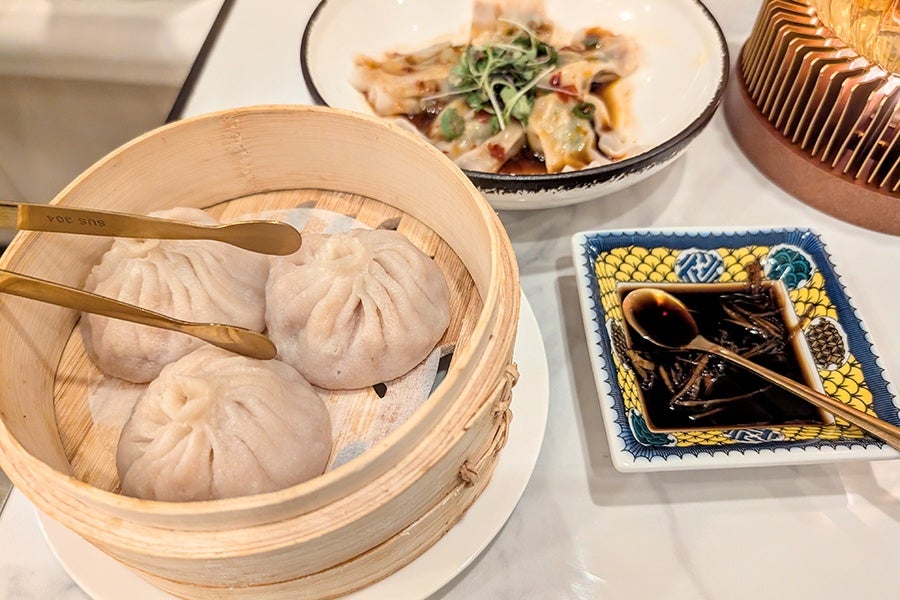 Three steamed dumplings in a bamboo steamer basket with gold tongs resting on top, accompanied by a bowl of dumplings in sauce garnished with microgreens and a small square dish of soy sauce with a gold spoon.