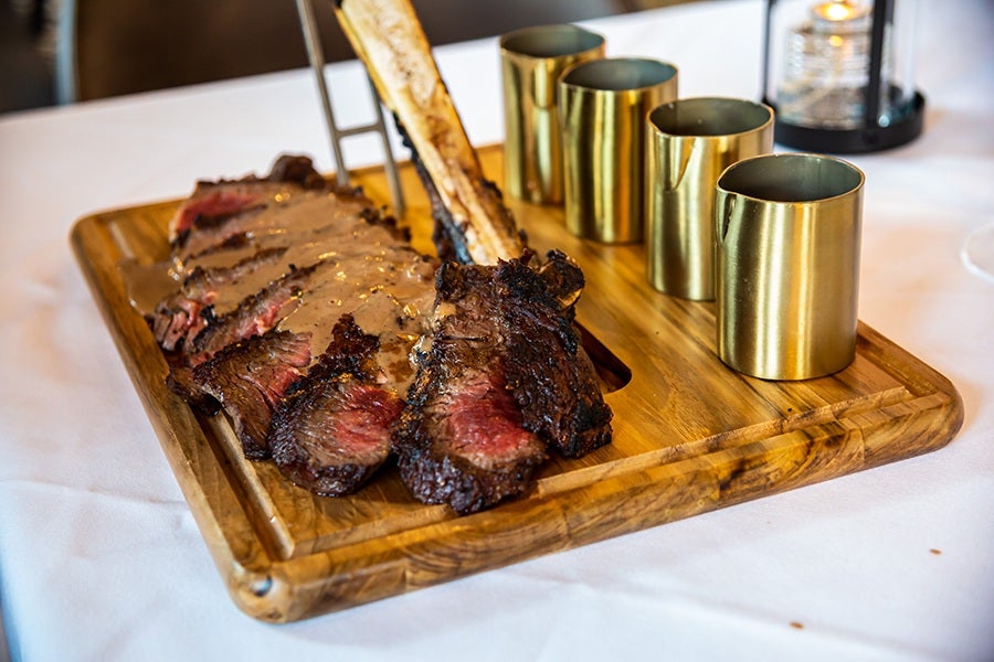 Sliced medium-rare steak with a bone, served on a wooden cutting board alongside four gold-colored metal cups. The steak is topped with a creamy sauce. The setting includes a white tablecloth and a candle lantern in the background.