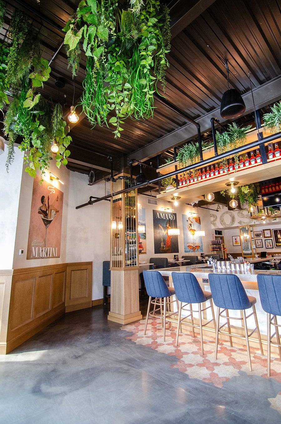 Modern bar interior with a polished concrete floor and patterned tile near the bar. The bar features a white marble countertop with blue upholstered bar stools with gold legs. Above the bar, there is a suspended shelf holding bottles and greenery. The ceiling is dark with hanging plants and exposed industrial lighting. The walls have framed posters and artwork, including a large "Martini" poster on the left. The space combines natural elements with contemporary design.