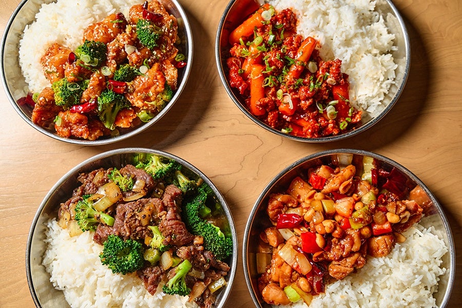 Four bowls of Asian-style dishes, each served with white rice. The top left bowl contains crispy chicken pieces with broccoli and dried red chilies. The top right bowl has a spicy dish with rice cakes and ground meat garnished with chopped green onions. The bottom left bowl features beef stir-fried with broccoli and onions. The bottom right bowl includes a mix of chicken, peanuts, diced red bell peppers, and celery in a savory sauce. All dishes are presented in round metal bowls on a wooden surface.