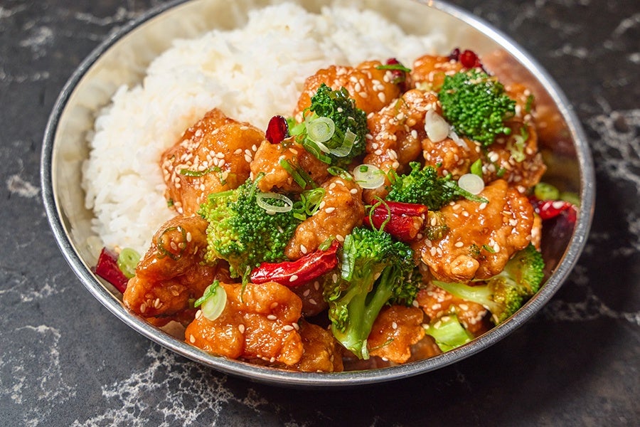 A bowl of white rice served with pieces of orange-glazed chicken, broccoli florets, red chili peppers, and sprinkled with sesame seeds and chopped green onions.