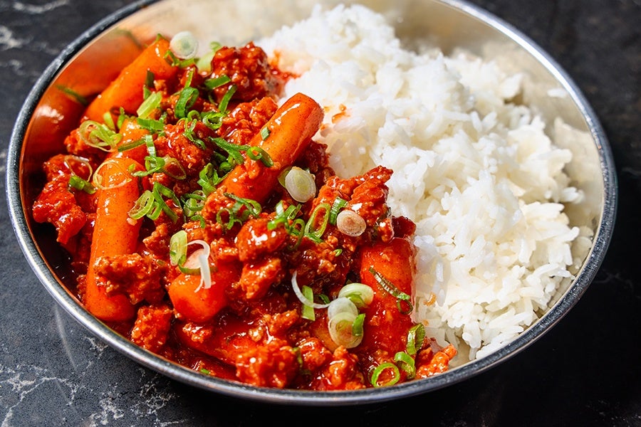 A metal bowl filled with white rice and a spicy red sauce dish containing ground meat, baby carrots, and chopped green onions sprinkled on top. The bowl is placed on a dark marble surface.