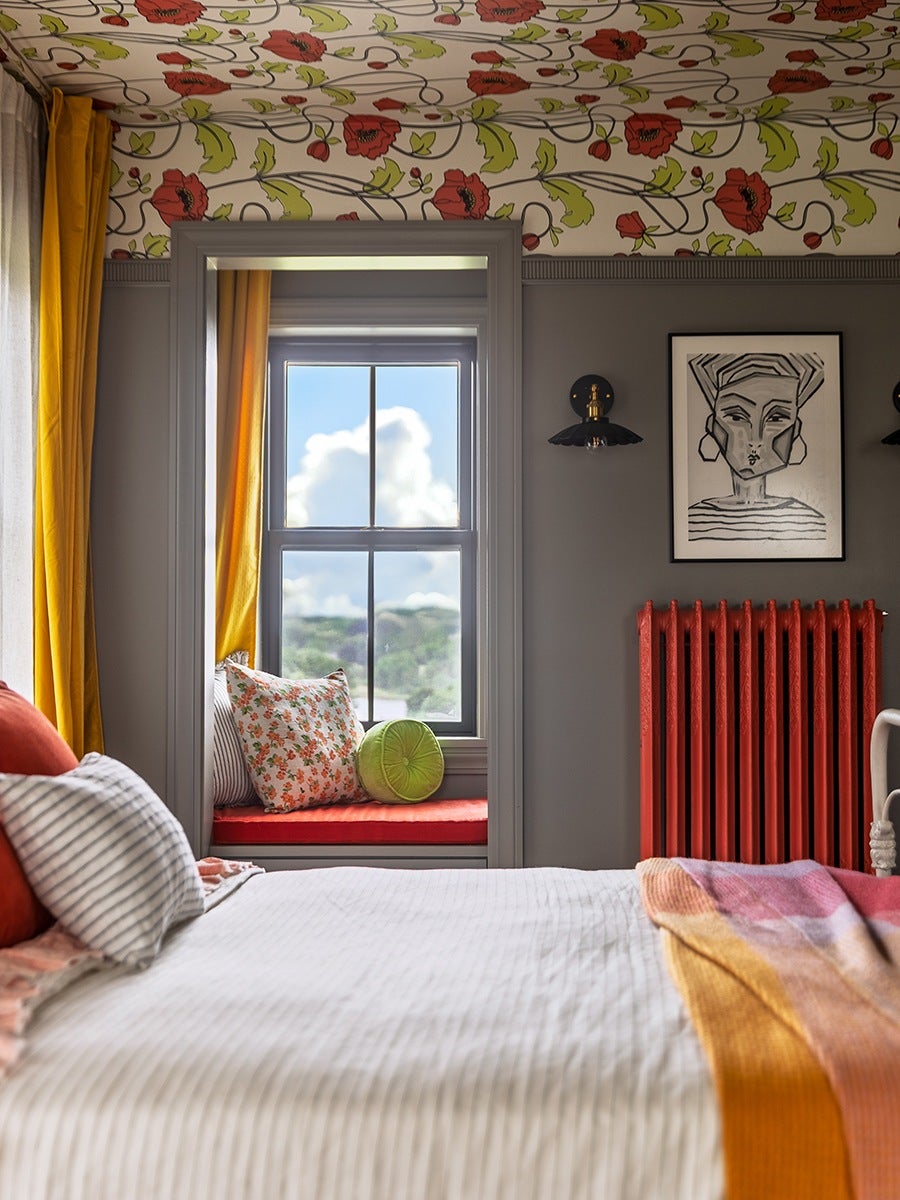 A cozy bedroom corner features a window seat with a red cushion and three pillows: one floral, one striped, and one round green. The walls are painted gray with a red radiator beneath a black and white abstract portrait. The ceiling and upper wall area are decorated with floral wallpaper in red, green, and black on a white background. Yellow curtains frame the window, and the bed in the foreground has white striped bedding with orange and pink blankets and pillows.