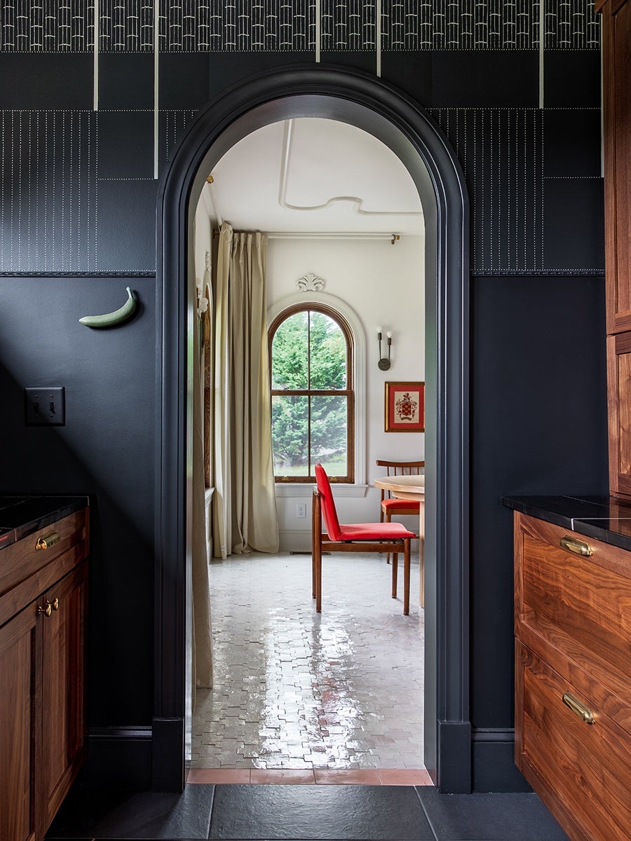 Arched doorway with a black frame leading from a dark kitchen with wooden cabinets and brass handles into a bright room featuring a red chair, a round wooden table, beige curtains, a tall arched window with a view of green trees, and a framed artwork on the wall. The floor in the bright room is light-colored and reflective.