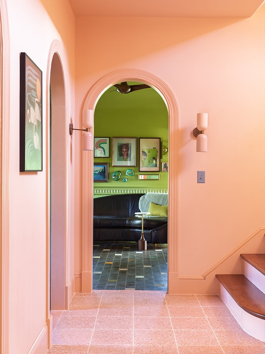 A hallway with peach-colored walls and tiled floor leads to a room with bright green walls. The hallway features an arched doorway, two wall sconces, a framed artwork on the left wall, and wooden stairs on the right. Inside the green room, there is a black leather sectional sofa with a yellow pillow and a white throw, a small round side table, and a gallery wall with various framed pictures and decorative items. The floor in the green room has dark, multicolored tiles.