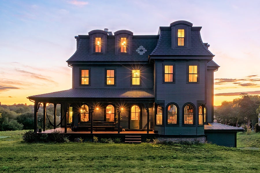 A large, dark-colored Victorian-style house with multiple windows lit from inside, featuring a wraparound porch with hanging lights and a hammock, set against a sunset sky and surrounded by green grass and trees.