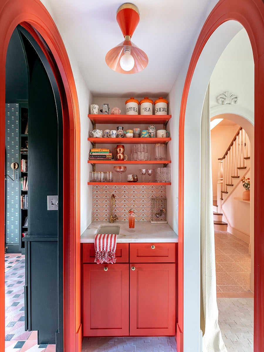 A small kitchenette with bright red cabinetry and matching red shelves holding mugs, glassware, and labeled canisters for coffee, tea, and sugar. The backsplash features a geometric tile pattern in soft pink and white. A brass faucet is installed over a small sink, and a red and white striped towel hangs from the cabinet. The space is framed by red arched doorways, with a red ceiling light fixture above. The adjacent rooms visible through the arches have contrasting dark and light color schemes.
