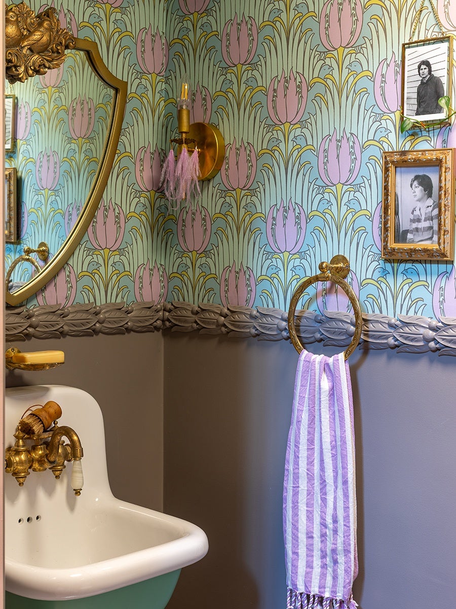 A vintage-style bathroom corner features a white sink with brass faucets and a wooden brush. Above the sink is a gold-framed mirror with an ornate top. The walls are decorated with pastel floral wallpaper in shades of green, pink, and blue, with a decorative molding border separating the wallpaper from the solid taupe lower wall. A brass towel ring holds a purple and white striped towel. Two framed black-and-white portraits hang on the wall, one in a gold ornate frame and the other in a simpler gold frame with a plant vine draped over it. A brass wall sconce with pink tassels is mounted above the sink.