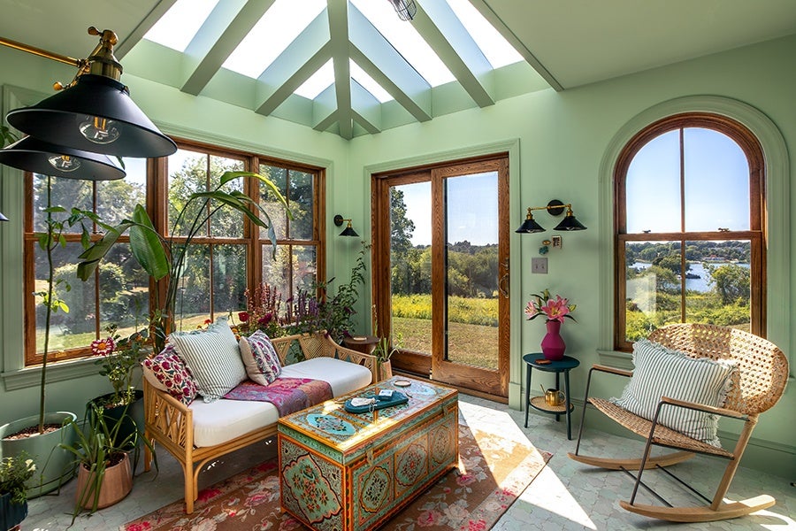Sunlit room with mint green walls and large wooden-framed windows, featuring a white cushioned wicker sofa with floral and striped pillows, a colorful ornate wooden chest as a coffee table, various potted plants, a wicker rocking chair with a striped pillow, and a small round side table with a pink vase of flowers. The ceiling has exposed beams with skylights, and the room overlooks a green landscape with trees and a body of water in the distance.
