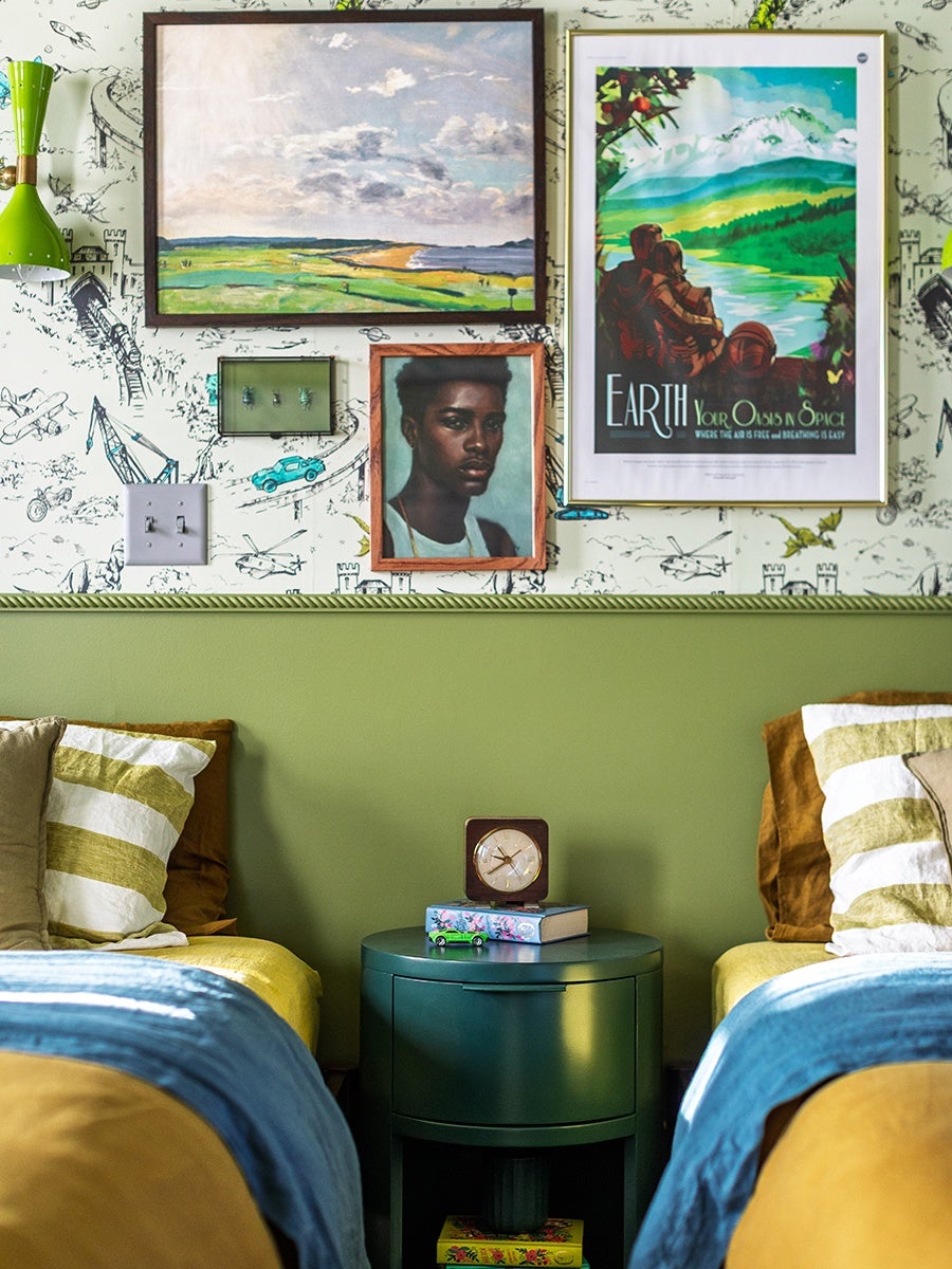 Two beds with striped pillows and blue blankets flank a dark green round nightstand. On the nightstand are a small clock, a book, and a toy car. The wall behind the beds is painted green on the lower half and features wallpaper with black and white sketches of various objects on the upper half. Above the beds are four framed pictures: a landscape painting, a portrait of a person, a green panel with hooks, and a colorful poster with the text "EARTH Your Oasis in Space." A green wall-mounted lamp is on the left side.