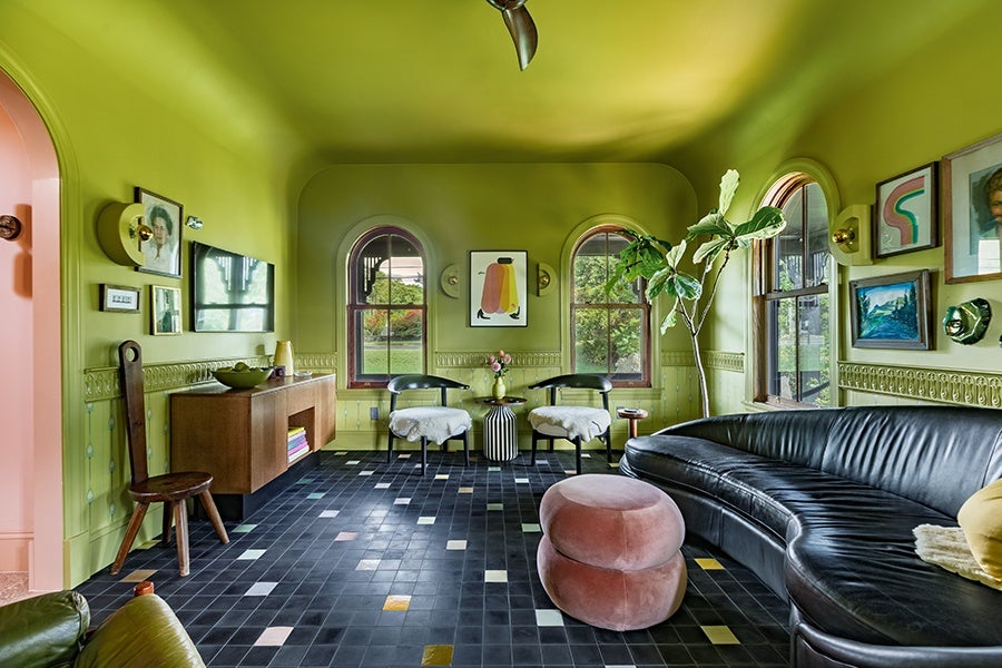 Living room with olive green walls and ceiling, featuring two arched windows. The room has a black leather curved sofa with a yellow pillow, two black chairs with white cushions, a striped round side table with flowers, and a pink double-stacked ottoman. The floor is tiled in black with scattered colored tiles. A wooden sideboard with books and decorative items is against one wall, which is adorned with various framed artworks and a large leafy plant near the windows.