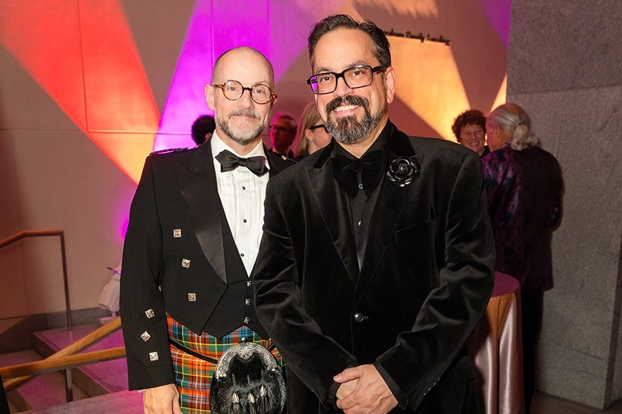 Two men dressed formally at an indoor event. The man on the left is wearing a black tuxedo jacket with silver buttons, a white dress shirt, a black bow tie, and a colorful tartan kilt. He also has round glasses and a bald head with a beard. The man on the right is wearing a black velvet suit jacket with a black shirt and a black bow tie, along with rectangular glasses and a beard. The background features colorful lighting with red, purple, and orange hues, and other people are visible in the background.