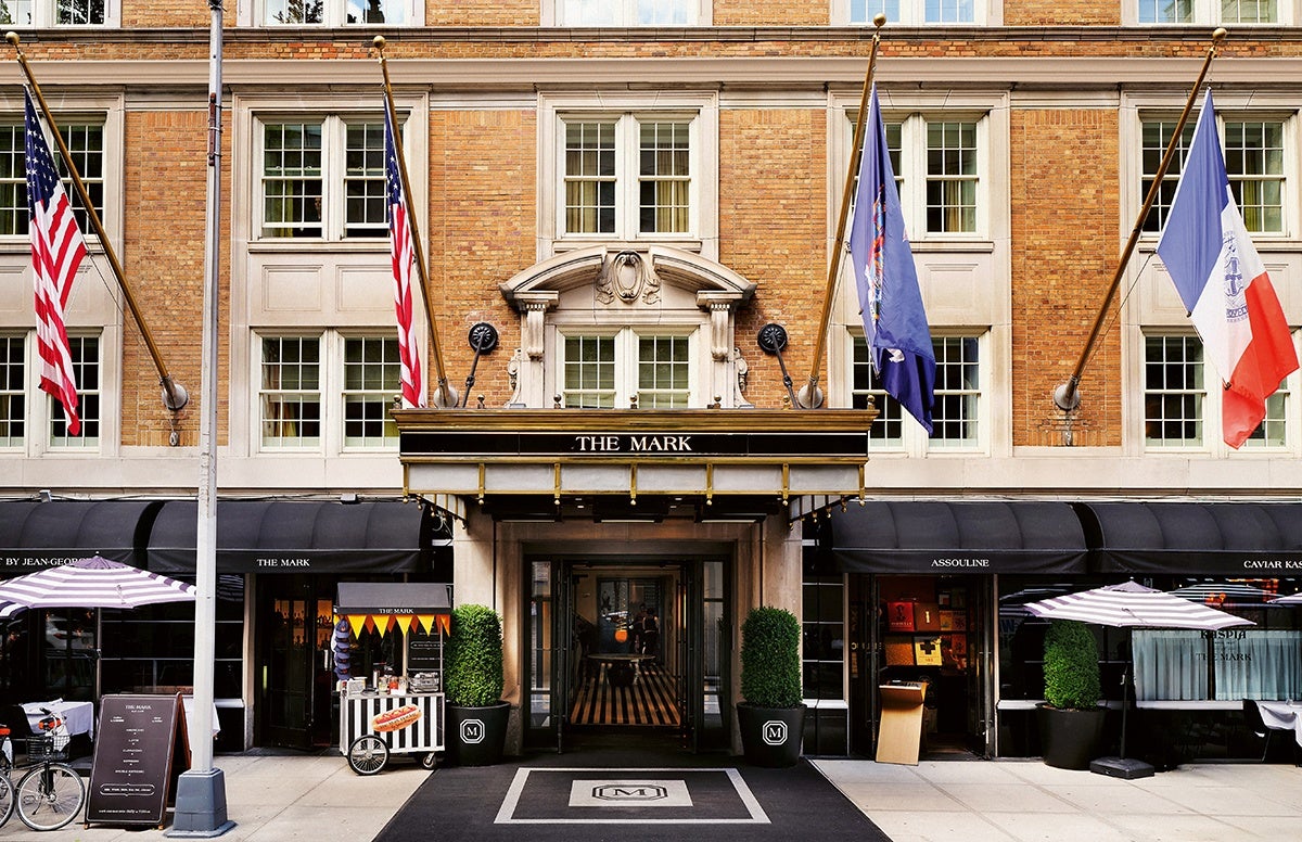 The entrance of The Mark hotel features a classic brick facade with large windows and a black and gold awning displaying "THE MARK." Four flags are mounted above the entrance, including two American flags, a New York state flag, and a French flag. Two potted green shrubs flank the doorway, and a black carpet with the hotel's logo leads inside. On either side of the entrance are black awnings for adjacent businesses, with outdoor seating areas shaded by striped umbrellas. A small cart with a hot dog sign is positioned near the left side of the entrance.