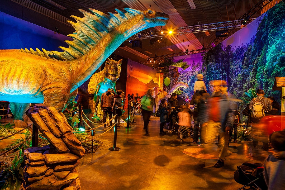 A brightly lit indoor dinosaur exhibit featuring large, detailed dinosaur models, including a prominent yellow and blue spiked dinosaur in the foreground and a T-Rex model behind it. The exhibit is decorated with artificial plants and rocks, with a backdrop of forest scenery. Several people, including children and adults, are walking around and observing the display, some appearing blurred due to motion. The setting has a warm, colorful lighting ambiance.