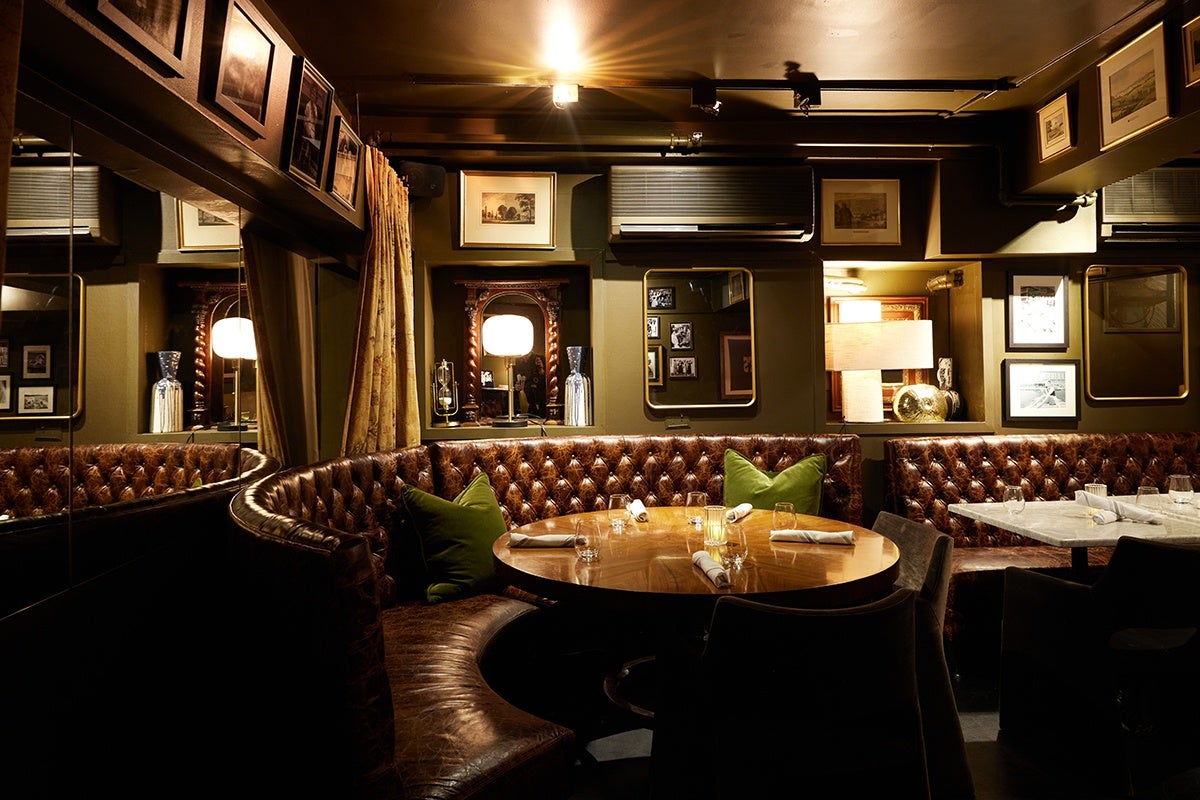 A cozy, dimly lit restaurant corner with a curved brown leather tufted banquette seating around a round wooden table set with napkins and glasses. The seating area includes green accent pillows. The walls are adorned with framed pictures and mirrors, and there is a warm table lamp and decorative vases in recessed wall niches. The overall ambiance is intimate and vintage.