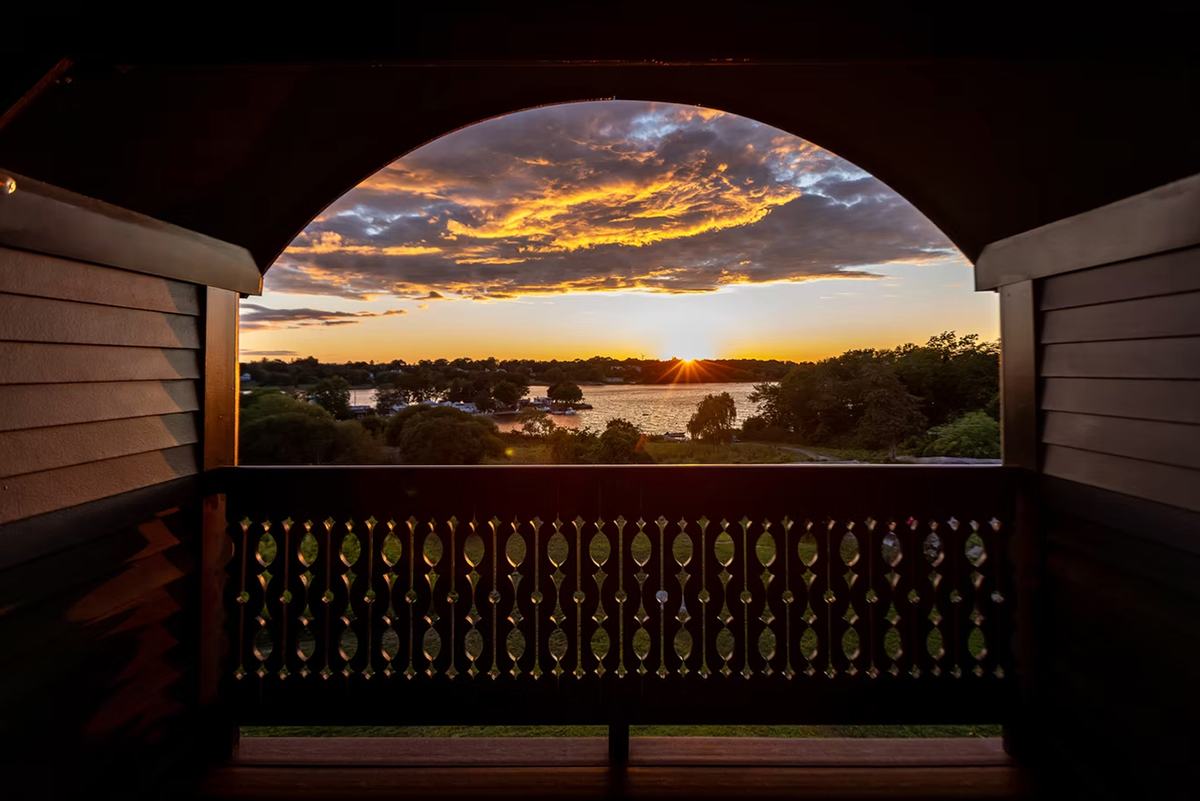 View of a sunset over a lake with trees and boats, framed by a dark wooden balcony with decorative railing and an arched opening. The sky features dramatic clouds illuminated with orange and yellow hues.