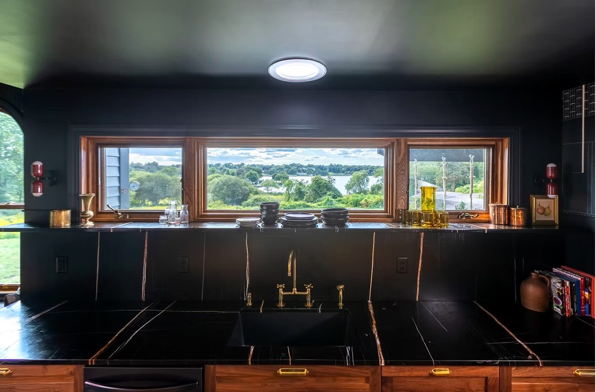 A modern kitchen sink area with black marble countertops and backsplash featuring subtle white veining. The sink is black with a gold faucet and handles. Above the sink is a wide wooden-framed window offering a scenic view of greenery and a body of water. The countertop holds various items including stacked black bowls and plates, glass bottles, a yellow glass pitcher with matching glasses, copper containers, a small framed picture, and a few books. The cabinetry below is wooden with gold handles. The ceiling and walls around the window are painted black, and there is a round ceiling light above the window.