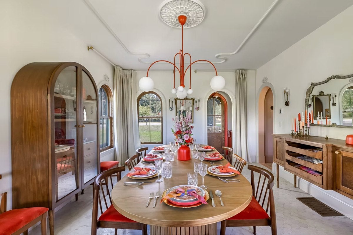 Dining room with a large oval wooden table set for eight, featuring colorful plates and napkins, crystal glassware, and a red vase with flowers as a centerpiece. The chairs have wooden frames with red cushions. A modern orange chandelier with white globes hangs above the table. The room includes a wooden cabinet with glass doors on the left and a wooden sideboard with candles and a large ornate mirror on the right. The walls are white with arched windows and doorways, and light curtains frame the windows.