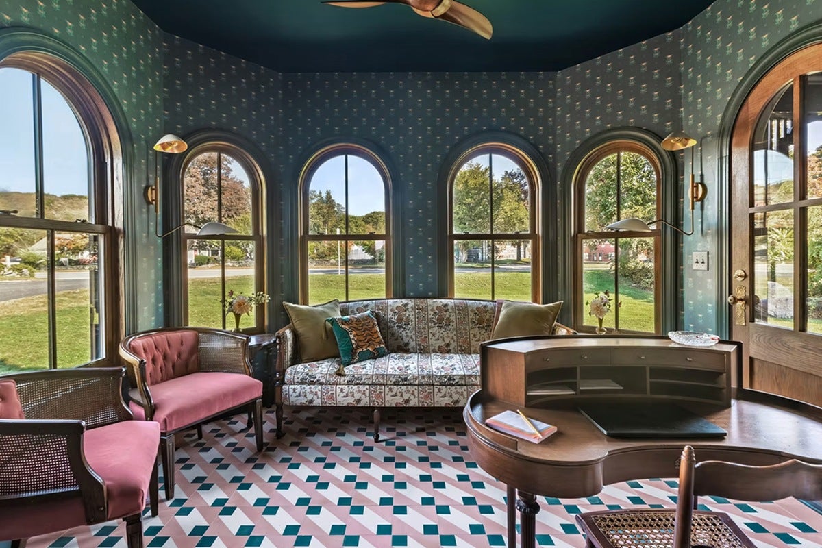 A cozy room with large arched windows featuring wooden frames, floral wallpaper, and a patterned tile floor. The room contains a floral upholstered sofa with green and patterned pillows, two pink cushioned chairs with wooden frames, a wooden curved desk with a notebook and pencil on it, and two small side tables with flower vases. Two wall-mounted lamps with brass fixtures flank the sofa, and a wooden door with glass panels is visible on the right.