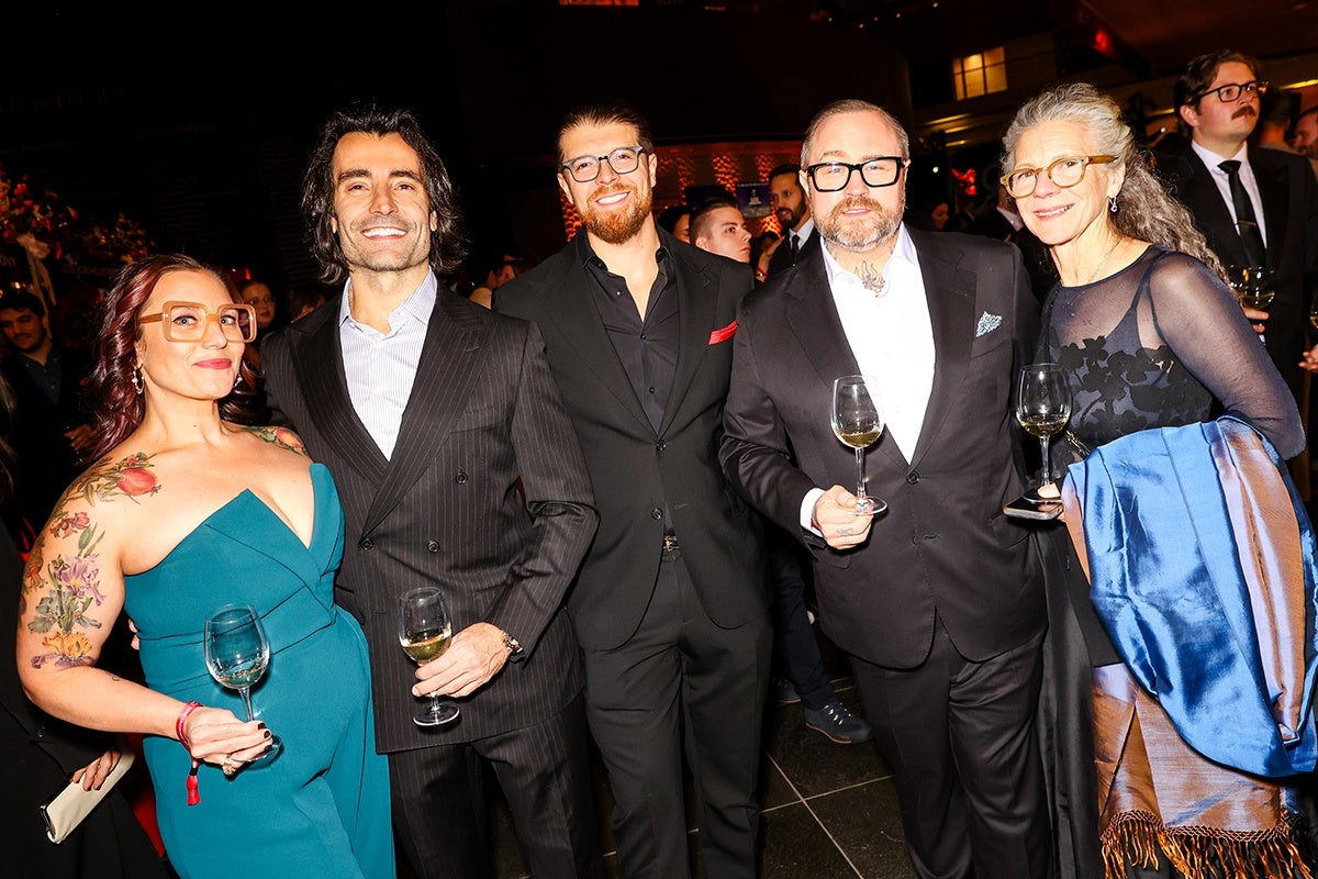Five people dressed in formal attire stand closely together, smiling and holding wine glasses. From left to right: a woman with red hair, large glasses, and colorful floral tattoos on her arm wears a teal dress; a man with dark hair and a pinstripe suit; a man with a beard and glasses in a black suit with a red pocket square; a man with a beard, glasses, and a tattoo on his neck in a black suit and white shirt; and a woman with long gray hair, glasses, a black dress with floral patterns, and a blue shawl draped over her arm. The background is dimly lit with other people visible.