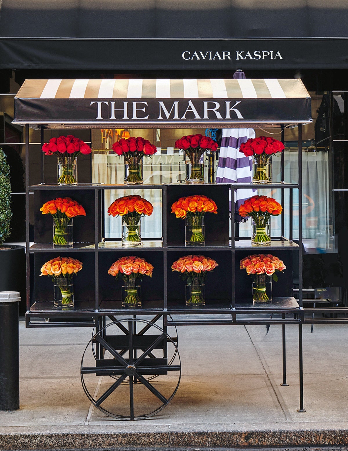 A black flower cart with a striped beige and white canopy labeled "THE MARK" displays twelve glass vases filled with roses. The top row features red roses, the middle row has orange roses, and the bottom row contains peach-colored roses. The cart has large black wheels and is positioned on a sidewalk in front of a building with a black awning that reads "CAVIAR KASPIA.