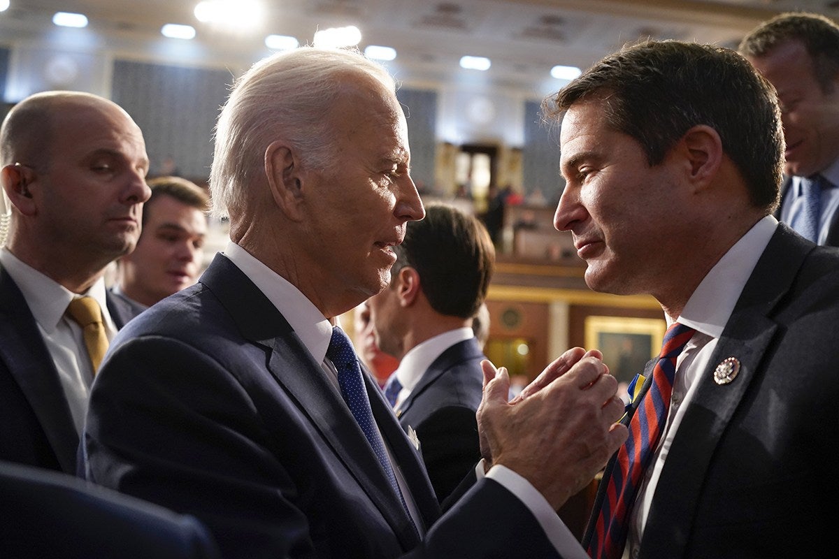 Two men in suits are engaged in a close, friendly handshake and conversation in a formal indoor setting with other people in the background. The man on the left has white hair and is wearing a dark suit with a white shirt and blue tie. The man on the right has dark hair and is wearing a dark suit with a white shirt and a red and blue striped tie. The background shows a well-lit room with blurred figures and framed artwork.