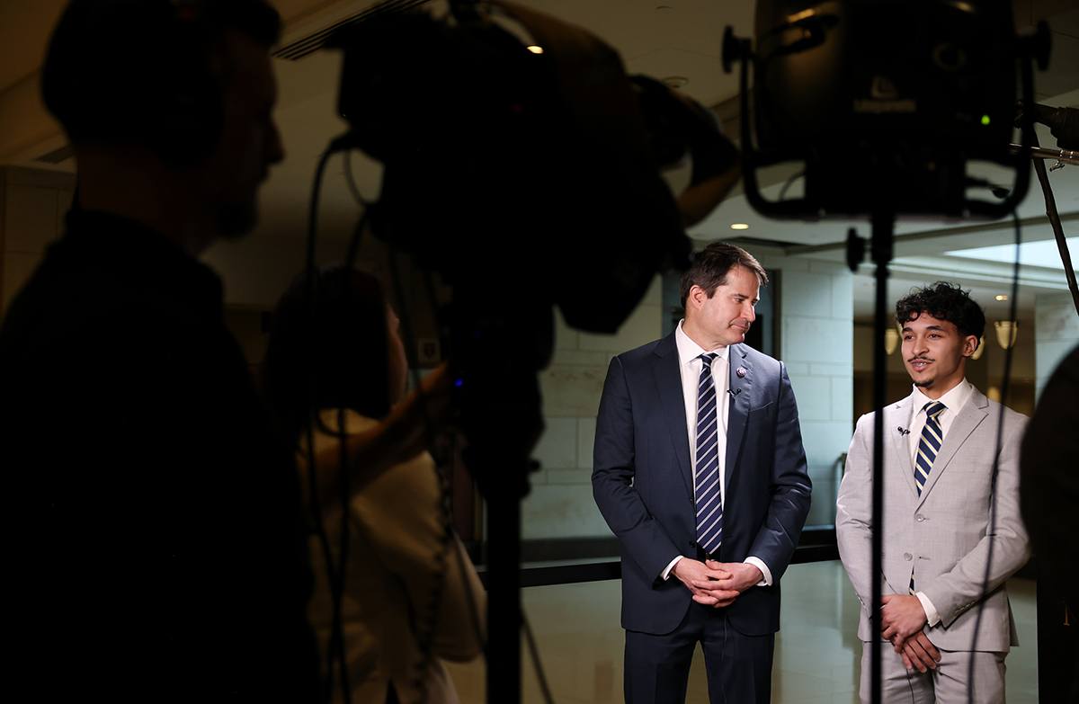Two men in suits stand side by side in a well-lit indoor setting, facing slightly towards each other. The man on the left wears a dark navy suit with a white shirt and a striped tie, while the man on the right wears a light gray suit with a white shirt and a similar striped tie. Both have microphones clipped to their lapels. In the foreground, there are silhouettes of camera equipment and crew members, indicating a filming or interview setup.