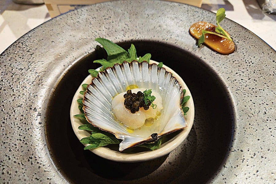A scallop dish served in a half shell placed on a small bowl with green leaves underneath. The scallop is topped with black caviar and finely chopped green garnish, with a drizzle of yellow oil. The bowl is set on a large, textured, dark gray plate with a small dollop of brown sauce and a green leaf garnish on the rim.