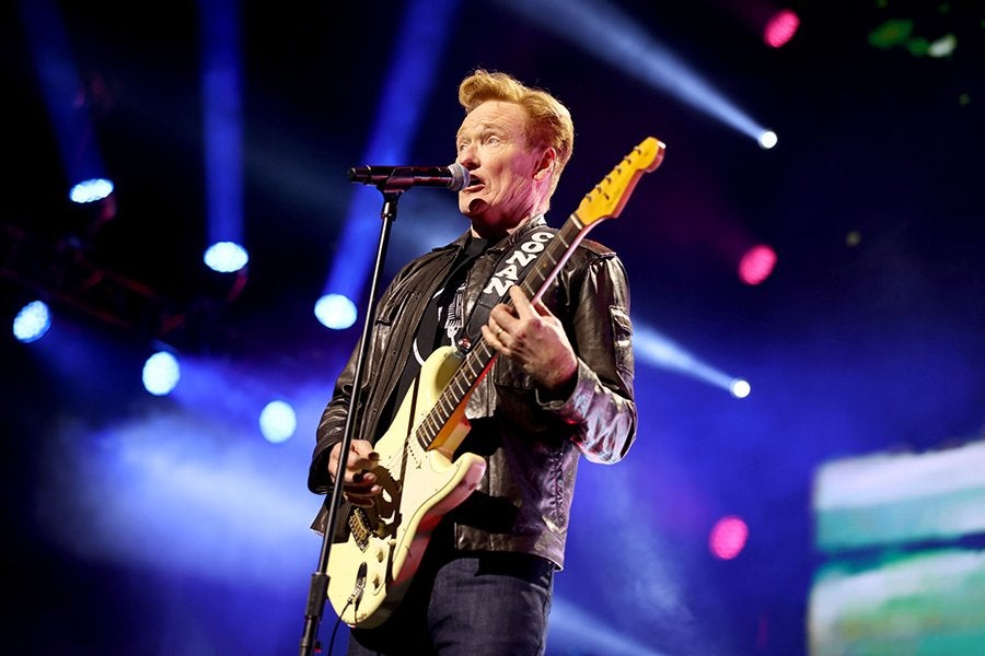 A male musician with light hair is performing on stage, playing a cream-colored electric guitar and singing into a microphone. He is wearing a black leather jacket and dark pants, with stage lights and smoke creating a vibrant background.