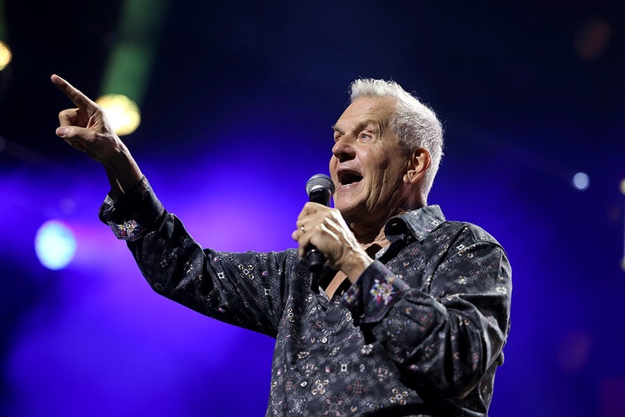 An older man with gray hair is singing or speaking into a microphone while pointing with his left hand. He is wearing a dark, patterned shirt and is illuminated by stage lights with a purple and blue background.