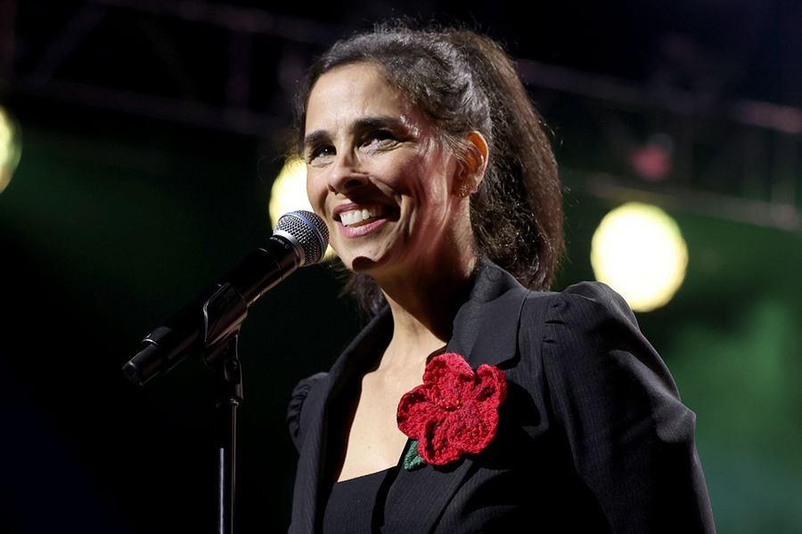 A woman with dark hair tied back in a ponytail is smiling while speaking into a microphone. She is wearing a black outfit with a large red flower accessory pinned to her chest. The background is dark with blurred lights.